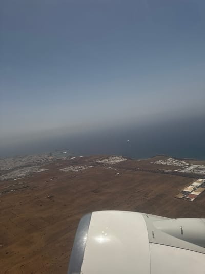 A landscape photograph taken from an aerial perspective from the window of a high altitude aircraft shows a land mass bordering a coastline, including a town in the municipality of San Bartolomé, Spain. The landmass is light brown, transitioning into a coastline with a few built structures near the coast. The horizon line separates the earth from the sky, which is light blue and grey. The right side of the image shows an aircraft wing.