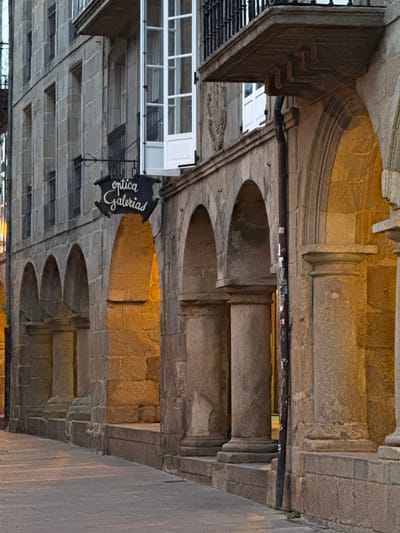 The image shows a view of the Rúa do Franco in Santiago de Compostela, Spain. It is an outdoor scene with a focus on the architecture of the buildings lining the street. The buildings are constructed from stone, with arched doorways and balconies. The light suggests it is either early morning or late evening, with a warm glow emanating from within some of the arched openings. A sign with the words "optica galerias" is visible hanging from one of the buildings. The street itself appears to be paved with stone. There are no people visible in the image. The overall impression is one of historical charm and a quiet urban streetscape.
