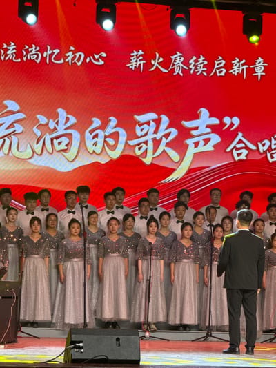 This image captures an indoor choir performance or rehearsal, likely taking place in Guta, China. The scene is dominated by a large red backdrop or screen displaying prominent white Chinese characters. The main text, in a large, stylized font, reads "流淌的歌声”合唱" which translates to "'Flowing Songs' Choir" or "Chorus." Above this, another line of text says "流淌忆初心 薪火赓续启新章," which can be interpreted as "Flowing, remembering the original aspiration; the torch is passed, starting a new chapter."
A large choir, comprising both men and women, is arranged in multiple rows, facing away from the viewer towards the backdrop. The women are dressed in elegant, light-colored, possibly silver or sequined gowns with long skirts, while the men wear white shirts, black bow ties, and black trousers. Many choir members have individual microphones or shared microphones positioned in front of them. In the foreground, with his back to the viewer, stands a conductor dressed in a dark suit, actively leading the choir.
The stage is illuminated by several spotlights, some casting bright white light and others a yellow-green hue, suggesting a performance setting. To the right of the choir, stage equipment such as large black speakers and cables are visible. The expressions of the choir members appear focused and engaged in the singing. The overall atmosphere suggests a formal and well-prepared musical event.