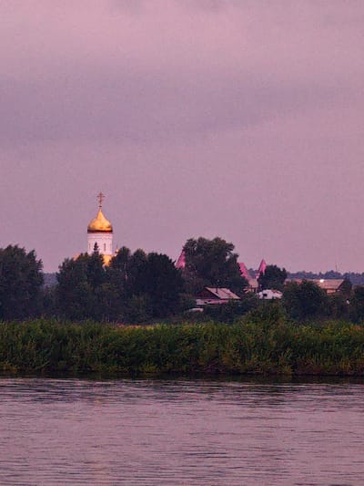 This media file depicts a serene landscape scene. The primary subjects are a river in the foreground and a village nestled among trees in the background. A prominent feature of the village is a white church with a glistening golden dome and a cross on top. Several other buildings, some with pink roofs, are visible, interspersed with lush green trees. The scene is bathed in the soft, hazy light of either dawn or dusk, indicated by the muted purple and pink hues of the sky. The water in the river has a gentle ripple, reflecting the diffused light. The overall atmosphere is peaceful and tranquil, suggesting a quiet rural setting in Kazachinskoe, Russia. There is no visible text in the image.