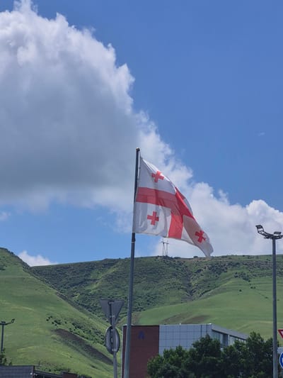 The image shows the flag of Georgia waving in the wind. In the background is a green mountain, partially obscuring a building with a red brick base and grey upper portion. There are utility poles and street signs in the vicinity of the flag. The sky is blue with some white clouds. The location is likely in or near Rustavi, Georgia, given the flag.