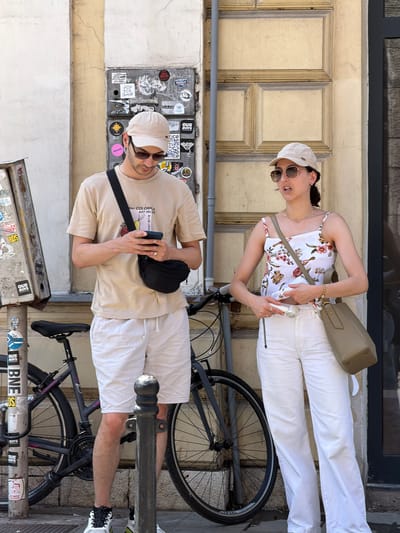 Two people, a man and a woman, are standing on a sidewalk next to a building in Sarajevo, Bosnia and Herzegovina. The man, wearing a light tan t-shirt, light beige shorts, a tan baseball cap, and sunglasses, is looking down at his phone. He has a black fanny pack slung across his chest. The woman, wearing a floral print camisole, white pants, a tan baseball cap, and sunglasses, is looking towards her right and speaking, with her mouth slightly open. She has a light tan crossbody bag. Between them, a black bicycle is leaning against a pole covered in stickers. The building behind them is light yellow with recessed panels and has a dark gray window or doorway to the right. The sidewalk appears to be paved with gray tiles. The overall lighting suggests it is daytime and sunny.