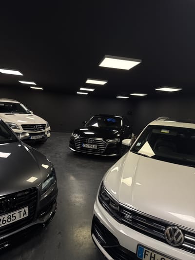Four cars are parked in an indoor garage with a black ceiling and dark gray walls. The garage has multiple bright, rectangular LED lights on the ceiling. From left to right, the cars are a dark gray Audi, a white Mercedes-Benz SUV, a black Audi sedan, and a white Volkswagen SUV. The dark gray Audi is in the foreground on the left, showing its front grille and license plate which reads "2685 TA". The white Mercedes-Benz is parked behind the dark gray Audi, with its front grille and license plate visible, reading "P 9978 MA". To the right of the Mercedes is a black Audi sedan, displaying its front grille and license plate "CB 7888 AO". A white Volkswagen SUV is positioned in the foreground on the right, with its front end partially visible, showing the grille and the Volkswagen logo. The license plate of the Volkswagen appears to be a European plate with a French flag symbol. The cars are clean and appear to be well-maintained. The floor is a dark, polished concrete. The overall impression is of a showroom or a high-end car storage facility. There are no people visible in the image. The location is in the city of Giurgiu, Romania.