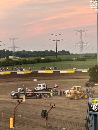 The image captures a dirt oval race track in Joliet, Illinois, during dusk. The sky is painted with soft pink and orange hues from the setting sun, with prominent power lines and transmission towers stretching across the horizon. In the foreground, a tow truck is parked on the dirt track, and nearby, a yellow front-end loader and a red ATV are also present. Several people are seen on and around the vehicles, some appearing to be working or observing the scene. A fence separates the track from a spectator area where a group of people are gathered. In the distance, lush green fields and trees form the backdrop, suggesting a rural or semi-rural setting on the outskirts of Joliet. A sign for "Dirt Oval Route 66" is visible on the right side of the image, identifying the location.