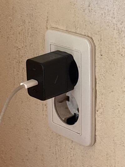 The image shows a close-up of a white electrical outlet on a textured, beige wall. A black power adapter, likely for a mobile device, is plugged into the top receptacle of the outlet. A white charging cable extends from the adapter. The bottom receptacle of the outlet is empty. The overall scene suggests a typical indoor setting, possibly a residential or hotel room in Krasnaya Polyana, Russia, given the outlet type which is common in the region.