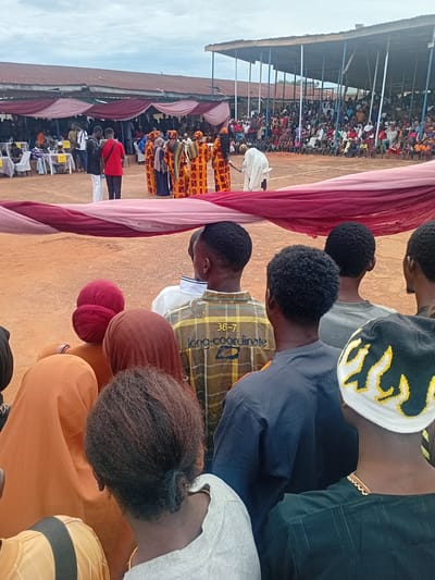 This photograph captures an outdoor public gathering or event in Ogbogbo, Nigeria. The foreground shows the backs of approximately eight individuals, observing the event. One person wears a brown and yellow plaid shirt with "36-7 long-coordinate" printed on the back. Another wears a dark shirt and a white beanie with yellow flame-like patterns. Several individuals have dark, curly hair; others wear head coverings in red, brown, and orange. A large, horizontally stretched drapery of pink and maroon fabric runs across the lower third of the image, acting as a decorative element or barrier.
In the midground, on an unpaved reddish-brown dirt surface, a group of approximately six individuals, primarily women, are dressed in vibrant, patterned traditional African attire, predominantly yellow, orange, and red. They appear to be performing or participating in a cultural ceremony. One individual in white robes is positioned near this group. To the far left midground, under a canopy structure, several tables and white plastic chairs are visible, some occupied. Two men stand conversing, one in a red shirt and the other in a white shirt.
The background features a densely packed crowd of spectators seated or standing on elevated bleachers, constructed with exposed metal poles. Behind these spectators, several low-rise buildings with rusted corrugated metal roofs are visible. The sky is overcast and uniformly gray.