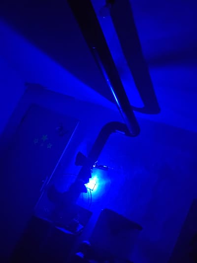 The image depicts an indoor scene illuminated by a bright blue light, casting strong shadows. In the foreground, a black oscillating fan is visible, angled upwards. The fan is positioned in front of a wall that appears to have some decorative elements, possibly glow-in-the-dark stars. To the left of the fan, a door is partially visible, with some indistinct objects attached to it. Above the fan and door, a curved pipe or conduit runs across the ceiling. The overall impression is that of a room with a dramatic, low-light ambiance.

The provided image does not contain any information that would allow for the determination of its location within the city of Daskotna, Bulgaria.