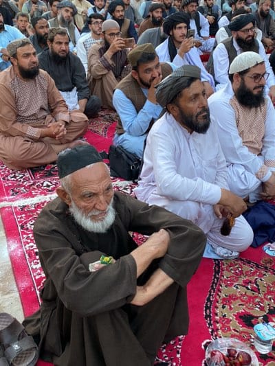 A medium shot captures a gathering of predominantly Pashtun men, seated on a red rug with a floral pattern, in what appears to be an outdoor urban setting in Kandahar, Afghanistan. In the foreground, an elderly man with a long white beard and a black cap is seated, wearing a dark brown tunic. His hands are clasped, and he holds prayer beads. To his right, another man in a white tunic and a grey turban is also seated, with his hands resting on his lap. Behind them, a crowd of men are visible, many wearing turbans of various colors, including black and white. Some men are looking forward, while others are engaged in conversation or looking at their phones. The scene suggests a communal event, possibly religious or political, given the attire and demeanor of the individuals. The background is blurred but suggests a city environment with buildings and other people. The overall atmosphere is one of quiet assembly.