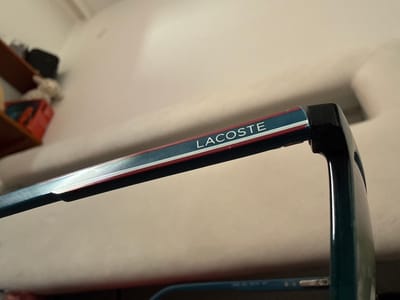 The image shows the arm of a pair of eyeglasses with the word "LACOSTE" printed on it in white letters against a blue and red stripe. The eyeglasses are dark green. The background is a light beige surface. A partially visible blurred object is located in the lower right corner of the image. The scene is not in Lüneburg, Germany.