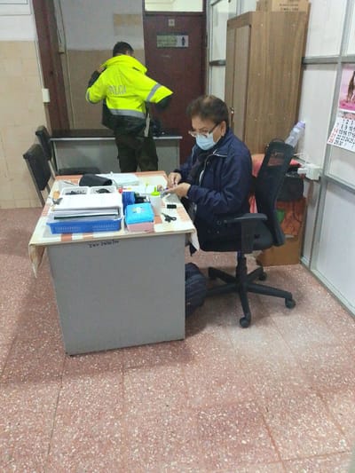 The image depicts an indoor scene, possibly an office or administrative area, in Santa Cruz de la Sierra, Bolivia. In the foreground, a woman wearing glasses and a face mask is seated at a desk, engaged in some activity, possibly administrative work. She is dressed in a dark jacket. The desk is cluttered with various items including papers, folders, a blue basket, and a small blue box. To the left of the desk, two black chairs are visible.

In the background, a person in a bright yellow reflective police jacket with "POLICIA" written on the back is standing and appears to be adjusting their clothing. This individual is wearing dark pants. Behind them, a wooden cabinet and a door with a restroom sign are discernable. The floor has a pinkish speckled terrazzo pattern. A calendar with a picture of animals is partially visible on a wall to the right. The overall environment suggests a functional but somewhat utilitarian space.