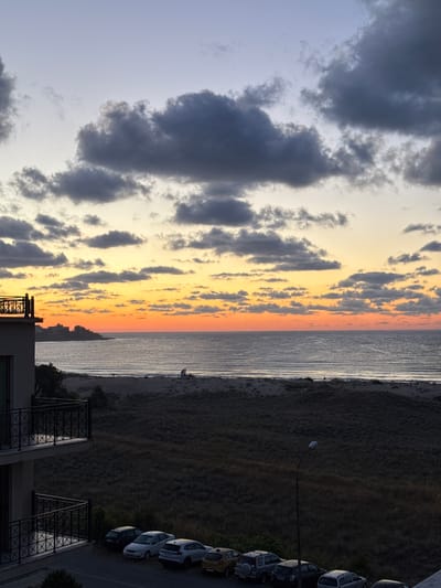 The image depicts a sunset over the Black Sea coastline near Primorsko, Bulgaria. A modern building with balconies is in the foreground, with parked cars on the ground level. Beyond the building is a sandy beach with vegetation, leading to the calm sea. The sky is a gradient of purple, orange, and yellow, with dark clouds scattered across it. The overall scene is tranquil, suggesting a peaceful evening on the coast.