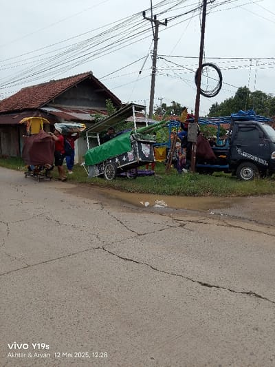 The image shows a street scene in Kecamatan Haurgeulis, Indonesia, located at the coordinates -6.419 latitude, 107.968 longitude. The scene includes a gray asphalt road with a large crack running down the center. To the left of the road, three people are walking. Two of them are men, and one is a woman. They are near a stall or food cart, similar in appearance to a hot dog stand. The cart has a green roof and a sign visible on the side. A black pickup truck with a white logo is parked at the edge of the street, on the right side of the frame. There is a person in the bed of the truck, facing away from the camera. The truck is parked near a wooden pole with a black electrical wire attached to it. The wire circles the pole. A second pole, with more electrical wires attached, runs along the left side of the street. The scene is under an overcast sky. The image also includes a house on the left side of the frame with a tile roof and a green garden area behind it. The background contains foliage.