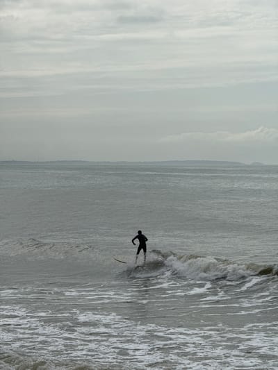 In a full shot of a surfer in Vietnam, a lone surfer is captured in the midst of riding a wave. The surfer, dressed in a dark wetsuit, stands on a surfboard that is partially submerged in the cresting wave. The surfer's body is angled as if in mid-motion, suggesting an active surf.
The scene unfolds against a muted backdrop. The ocean stretches to the horizon, its surface rendered in various shades of grey and white due to choppy waves and foam. The sky above is overcast, with layered clouds that create a diffused, soft light, contributing to the somber atmosphere. The distant shoreline is barely visible, appearing as a faint, dark line against the hazy sea and sky, possibly indicating the coast of Phan Thiết. The overall impression is one of a solitary individual engaged in a water sport on a cool, overcast day in Vietnam.
