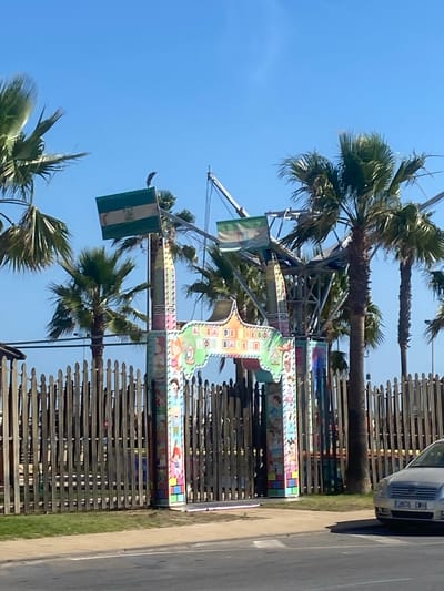 In Conil De La Frontera, Spain, a temporary amusement ride, likely a fairground attraction, stands under a clear blue sky. The ride's entrance features vibrant, cartoonish decorations and Spanish text that reads "DIVERION SIN FIN" (Endless Fun). Two flags, one with green and white stripes and a coat of arms, and another smaller flag with a similar design, are attached to the ride's structure. Palm trees surround the attraction, with one palm tree in the foreground showing signs of distress. A wooden fence encloses the amusement area, and a silver car is parked to the right of the frame, partially obscuring the fence and the ride. The ground in front of the fence is covered in green grass and a paved area, suggesting a public space adjacent to a road.
