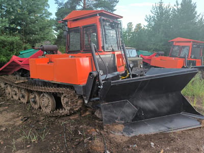 The image depicts an orange tracked vehicle with a large black plow attached to the front, positioned on a muddy, dirt terrain. The vehicle appears to be a specialized piece of equipment, possibly for forestry or construction, with a reinforced cab and visible tracks. In the background, other similar orange vehicles and a green and red attachment are visible, along with a military-style truck and dense coniferous trees. The setting suggests an outdoor, possibly rural or industrial area. The sky is overcast, indicating a temperate climate. There are no people visible in the image.