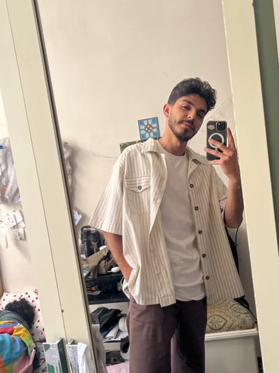 A young man takes a mirror selfie, captured from the chest up in what appears to be a domestic setting within As-Salihiya Municipality, Syria. He has dark, curly hair, a short beard, and a mustache, with a neutral expression. He is holding a dark-colored smartphone, likely an iPhone, distinguished by its camera module and a visible MagSafe ring on the back. He is dressed in a white t-shirt worn under an unbuttoned, light beige or cream-colored striped shirt with brown buttons, and dark brown trousers.
The reflection reveals a light-colored wall behind him, topped by a darker green or grey trim or shelf. A small, decorative square tile with a blue and brown geometric pattern is visible on the wall to the man's left. Below the mirror, a cluttered shelf or dresser contains various items including folded clothes, a pillow with a light blue and gold paisley pattern, white sneakers, and other personal belongings.
In the bottom right of the reflected scene, the head of another person with dark, curly hair is visible, resting on a white pillow adorned with red hearts and partially covered by a colorful, multi-patterned blanket, indicating they are asleep. The lighting is bright, suggesting it is daytime. The room presents a lived-in, somewhat cluttered appearance. No discernible text is visible in the image.