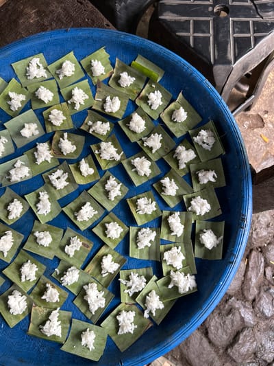 In the city of Manggis, Indonesia, a blue plastic tray is filled with small squares of green banana leaves, each topped with a portion of white rice. The tray is placed on a rough, outdoor surface, possibly ground or cobblestones, with some wooden elements visible at the edges of the frame. In the background, a black, textured object with grid-like markings appears to be a piece of furniture or equipment. The rice is arranged in small mounds, suggesting it is prepared for distribution or a ritual. The scene is captured from a high angle, looking down into the tray.