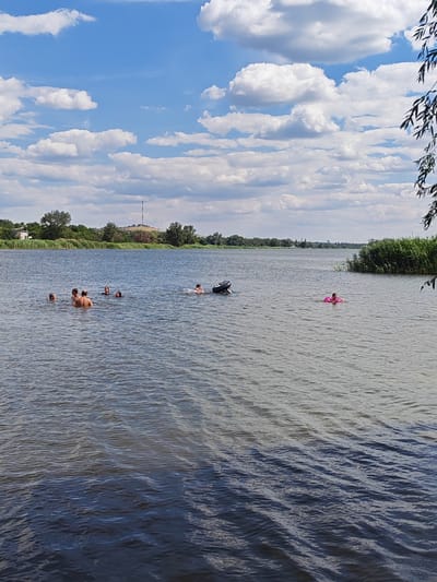 The image captures a wide, calm river with a group of people swimming in the water. The sky is bright blue with scattered white clouds. On the left bank of the river, there are trees and a building. In the background, a radio tower is visible on a hill. On the right bank, there is dense vegetation. One person is floating on a pink ring, and another is near a large tire in the water. The scene appears to be a recreational area within or near the city of Belaya Kalitva, Russia, likely a natural setting for swimming and leisure on the river.
