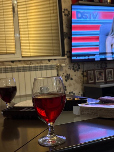 The image depicts an indoor residential setting in Cherven Bryag, Bulgaria, characterized by dim ambient lighting. In the foreground, positioned on a dark, reflective table surface, is a clear, stemmed glass containing a dark red liquid, likely wine. Condensation or small water droplets are visible on the exterior surface of this glass. To the left and slightly behind, a second, similarly filled stemmed glass is partially visible on the same table.

Behind the foreground glass, a black rectangular tray or container is present, alongside a partially visible white object and a cardboard box with visible text on its side.

The background features a window with closed light-colored horizontal blinds on the left. A white central heating radiator is mounted below the window. On the right, a flat-screen television is displaying content with the "DSTV" logo, characterized by horizontal red and blue graphical bands, and a partial view of a person. The television is mounted on a wall covered with patterned wallpaper, dark in tone with lighter floral or abstract motifs. Below the television, a dark piece of furniture holds three framed pictures arranged horizontally. No individuals are present within the visible frame. The overall scene suggests a prepared environment for relaxation or entertainment.