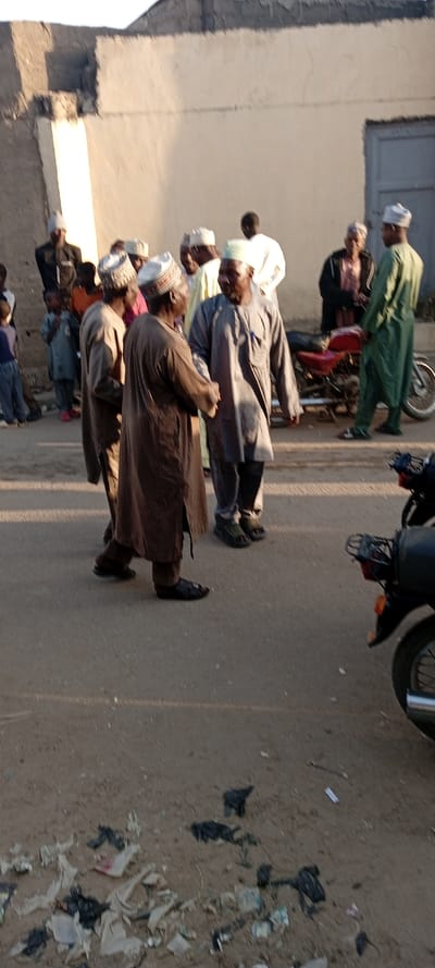The image captures an outdoor street scene in Kabuga, Nigeria, under bright daylight conditions, evidenced by clear shadows. Approximately 15 individuals are present, consisting primarily of adult males along with a few children. Most adults wear traditional Nigerian attire, including long robes (brown, grey, green, white, and beige) and various caps. In the foreground, two men in robes and caps stand facing each other, one in brown and one in grey, seemingly engaged in conversation. To their right, a man in a green robe and cap stands near a red motorcycle parked by a building. Another dark-colored motorcycle is partially visible in the bottom right corner. In the background, a light-colored, textured building wall with a dark doorway is visible. The ground surface in the foreground is a paved area, heavily scattered with white and black plastic bags, paper, and other debris.