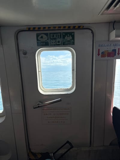 The image shows the interior of a ferry cabin, looking out through a door with a window. The door is made of gray metal and has various signs and markings. On the left side of the door, there are green and white signs indicating "EXIT" and "ASSEMBLY STATION," with symbols for people and a life raft. The window in the door looks out onto a bright, sunny seascape with blue water and white clouds. A metal handle is visible on the door, and a sign in Thai and English above it reads, "THE DOOR OF THE EMERGENCY EXIT MUST BE SHUT WHILE SAILING" and "PLEASE DO NOT OPEN THE CABIN DOOR DURING VOYAGE." Another sign above the door reads "BAGGAGE AREA" with illustrations of luggage and arrows. The overall impression is a functional, utilitarian space, likely on a ferry traveling through calm seas during the daytime. The lighting suggests it is daytime, and the clear sky indicates fair weather.
