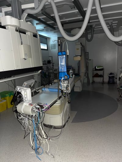 The image displays an unoccupied medical interventional room located in Dumbéa, France. The room features a light grey, speckled floor with a prominent darker grey semicircular pattern, light-colored walls, and a ceiling equipped with multiple corrugated grey ventilation ducts and recessed lighting fixtures.
A large, white medical procedure table, topped with a blue disposable sheet, dominates the central foreground. Attached to and hanging from the table's end is a dense cluster of white, blue, and black medical cables, tubes, and a white cylindrical apparatus. To the immediate left of the table, two open medical waste bins, one green and one yellow, are visible.
A large, white, rectangular medical imaging system is suspended from the ceiling and positioned over the left side of the procedure table. Further into the room, on the right, a second ceiling-mounted medical imaging unit, branded "PHILIPS" and "SLIDE," is present. An IV pole stands between the procedure table and the right imaging unit, supporting a large blue pressure bag cover and a smaller clear IV fluid bag.
In the background, a window with horizontal blinds and an air vent is visible. Multiple medical equipment carts, loaded with various devices, are situated near the window. On the far right, a three-tiered mobile shelf unit holds a black and green medical bag, and a blue container rests on the floor nearby. No individuals are present in the scene.