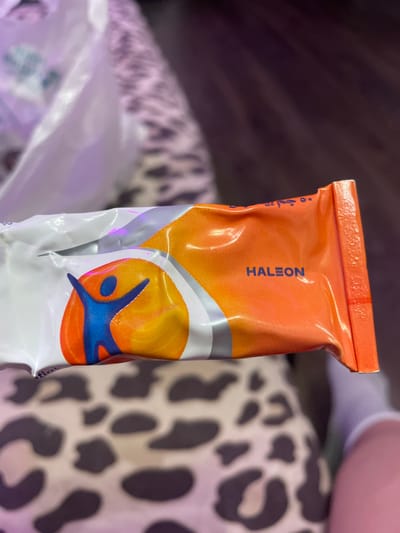 The image shows a close-up of a product packaging, which is primarily orange and white with blue and yellow accents. A blue human-like figure with outstretched arms is depicted on the white portion, set against a yellow and orange circular background. The word "HALEON" is clearly visible in blue text on the orange section of the packaging. The product is held at an angle, with the right edge of the packaging folded over, revealing a textured orange border. In the background, a person's legs are visible, wearing white socks and a light-colored garment with a leopard-like print. A portion of a white plastic bag is also visible in the upper left corner. The scene appears to be indoors, with the lighting suggesting it's not daytime. There is no indication of Vaganovo, Russia, or any specific city in the image.
