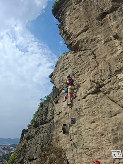 In Děčín, Czechia, two individuals, an adult and a child, are rock climbing a sheer cliff face equipped with a metal ladder and handholds. The adult, wearing a helmet, backpack, and climbing attire, is positioned above the child. The child, also wearing a helmet and a harness, is climbing below the adult. The rock face is a mix of rough, textured stone and some smoother sections, with visible bolts and metal rungs integrated into the surface for climbing. The background shows a bright blue sky with scattered white clouds, and a distant view of a town with buildings and some vegetation. A sign is visible on the rock face with text that appears to be in Czech, indicating a location or route.