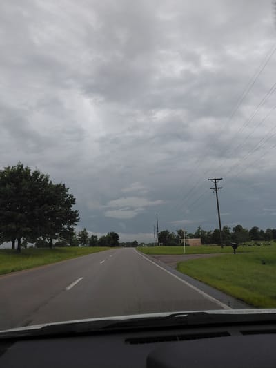The image depicts a roadside scene in Paris, United States, likely during the daytime. The perspective suggests the photo was taken from inside a vehicle, looking out. The primary subject is a road with white lane markings, curving gently to the right. To the left of the road, a grassy area is visible, bordered by a line of trees. In the distance, more trees and what appear to be gravestones suggest the presence of a cemetery.
Dominating the upper portion of the image is a cloudy sky. The clouds are grey and somewhat dense, indicating overcast or potentially stormy weather. There are several utility poles with power lines stretching across the frame, further contributing to the sense of a rural or semi-rural setting. One prominent pole with crossbars is centrally located. A sign is partially visible in the middle ground, but its text is not legible.
The overall mood of the image is somewhat subdued, likely due to the cloudy sky. The scene is peaceful and ordinary, capturing a moment of travel along a quiet road. The time of day is not definitively clear, but the ambient light suggests it is not dawn or dusk. The presence of the road, grass, trees, and utility poles provides context for a typical American landscape.