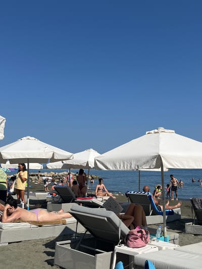 A sunny, clear blue sky dominates the upper portion of the image, overlooking a crowded beach in Germasogeia Municipality, Cyprus. White beach umbrellas provide shade for loungers on a dark, pebbly shore. Several people are visible, some relaxing on loungers, others wading or playing in the calm blue sea. A woman in a yellow top and dark shorts stands near an umbrella, while other individuals are scattered across the sand and in the water. In the foreground, a person lies on a lounger wearing a light purple bikini bottom. The overall scene depicts a lively summer day at the beach.