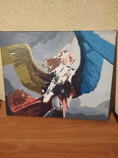 The image depicts a painting on a canvas. The painting features a central figure, possibly a young woman, with wings. She has brown hair and is wearing a pink floral headpiece and a white garment with dark accents. To her left, a large wing is depicted in shades of yellow and brown, while to her right, another wing is shown in vibrant blue. Below the figure, there is a large area painted in red and brown, which could represent a cloak or another wing. The background of the painting is a blend of grays and blues, suggesting a sky or mountainous landscape. The painting is placed on a wooden surface, likely a table or shelf, in front of a textured, light-colored wall. The painting itself is a work of art, with visible brushstrokes and areas that appear to be part of a paint-by-numbers kit, indicated by faint numbers within some of the colored sections. There is no indication of the city of Kamianske, Ukraine, in the image.