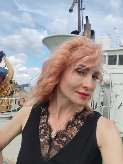 A woman with pink-tinted hair, wearing a black top with a lace insert and a silver necklace, poses for a selfie on a boat. She is looking at the camera with a slight smile, her lips painted red. In the background, a crane and the side of a white ship are visible against a cloudy blue sky. The location of the image is the city of Vasil'ki, Russia.
