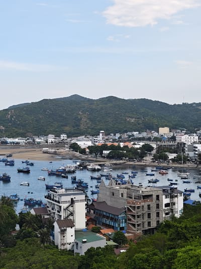 This is an aerial, full-frame, eye-level, vertical shot of the city of Vĩnh Hải, Vietnam, on a hazy day. The image captures a bay filled with numerous small, colorful fishing boats, their hulls varying in shades of blue, red, and yellow. Some boats are moored together, while others are scattered across the water. Beyond the bay, the city unfolds with a mix of buildings. In the foreground, there are a few trees and buildings, including a white multi-story structure with an external staircase. Further back, various buildings, some appearing to be under construction with exposed concrete and scaffolding, are visible. The urban landscape extends towards a lush, green, mountainous backdrop. The sky above is a pale blue with scattered white clouds. The scene depicts a bustling coastal town with a strong connection to its maritime activities.