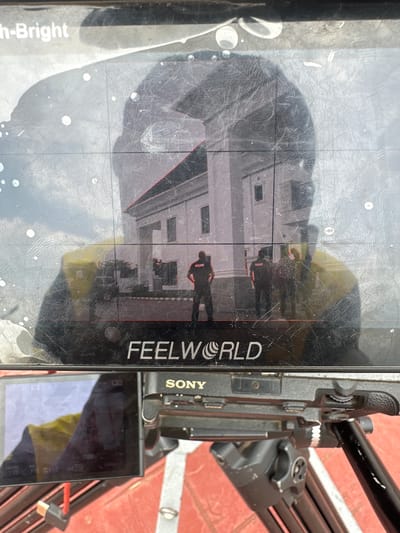 The image shows a close-up of a black monitor screen with a reflection of a building and several people. The monitor displays the brand "FEELWORLD" and "SONY" is visible on the camera below. The building appears to be a light-colored, possibly white, structure with windows and a distinct architectural style. In the reflection, several individuals are visible standing in front of the building. Some appear to be wearing dark uniforms with "POLICE" visible on the back of one. The overall scene suggests a filming or photography setup, possibly documenting an event or location. The environment outside the immediate focus appears to be outdoors with a cloudy sky reflected in the monitor. The location in Pakuro, Nigeria is not explicitly identifiable from the image itself, but the presence of police uniforms might suggest a public or official area. The monitor screen has numerous scuff marks and some circular smudges, indicating it might be used in various conditions. The camera below is mounted on a tripod, further emphasizing the professional recording context.