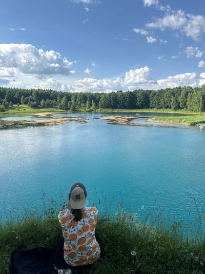 The image captures a scenic view of a bright turquoise lake surrounded by lush green trees and a blue sky with scattered white clouds. In the foreground, a person is seen from behind, sitting on a grassy embankment and looking out at the lake. The person is wearing a cap and an orange-patterned shirt. Several individuals are visible on the lake, some on paddleboards and others in a small boat, enjoying the water. The overall impression is one of a tranquil summer day in a natural setting. The location is identified as the city of Grudcino, Russia.