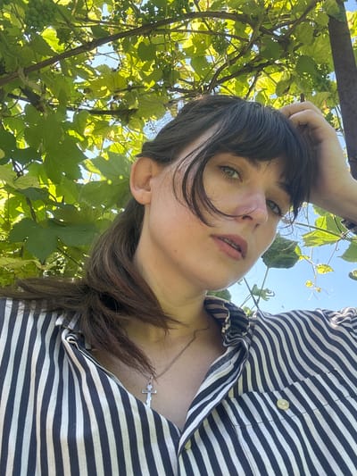 A low-angle, close-up selfie captures a young woman with dark brown hair and light green eyes, positioned in the foreground. She is outdoors, with lush green grape leaves and unripe green grapes arching over her head, set against a bright blue sky. She wears a black and white striped button-down shirt and a silver cross necklace. Her right hand is raised, resting on her forehead, and her gaze is directed slightly upward and to the left of the camera. The image appears to be taken in Zvonarevka, Russia, as indicated by the presence of grapevines, a common agricultural feature in the region.