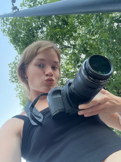 An individual is holding a Canon camera, with the lens facing the viewer. The person's mouth is pursed, and their eyes are looking at the camera. They are wearing a black tank top and a camera strap. In the background, there are green trees and a blue sky. The scene appears to be outdoors, possibly in Gošići, Montenegro.