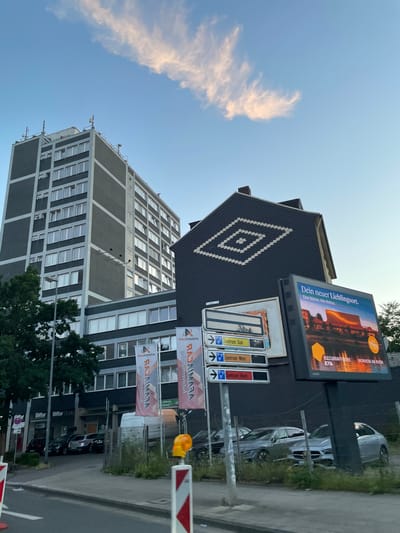 The image shows a street scene in Essen, Germany, with a tall, dark grey building on the left and a darker, gabled building on the right. A large billboard is attached to the gabled building, displaying an advertisement for "Dein neuer Lieblingsort" (Your new favorite place) with an image of a modern building with illuminated windows and text below referencing "KULTURRAFFINERIE MONHEIM AM RHEIN". Several directional signs are mounted on a pole in front of the billboard, indicating directions to "Zentrum Süd" (South Center) and "Zentrum West" (West Center). The street in the foreground is asphalt, with a sidewalk and some grassy areas. Several cars are parked behind a fence in front of the gabled building. The sky above is a clear blue with a wispy cloud. Banners with a red and orange design and the logo "RAJAMAIRA" are displayed on the left side of the frame, in front of the tall building. The tall building has a facade of alternating grey and white bands, with rows of windows. The gabled building has a dark, patterned roof.