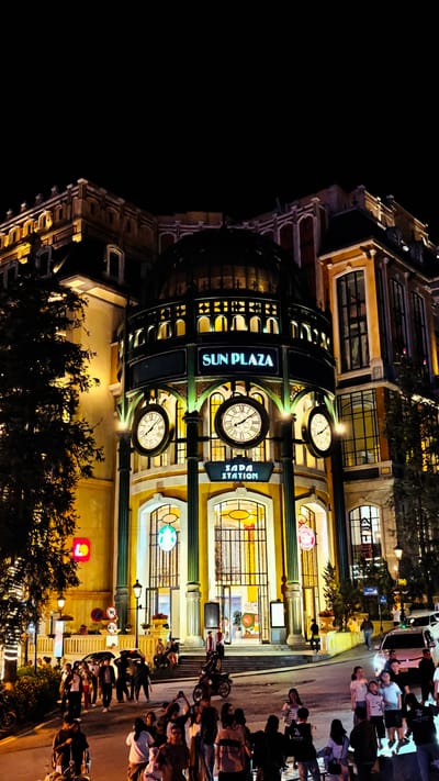 The image shows the Sun Plaza and Sapa Station in Sa Pa Ward, Vietnam, at night. The building is illuminated with lights, and there are three large clocks on the facade. People are walking on the street in front of the building. There are parked motorcycles and cars on the side of the road. The overall environment is urban, with trees visible on the left and right sides of the building.