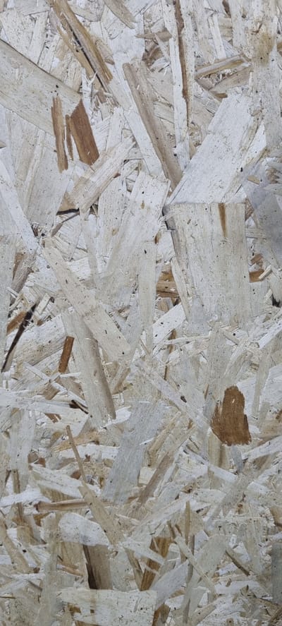 The image is a close-up, full-frame shot of oriented strand board (OSB). The texture is rough and uneven, composed of numerous wood chips and strands of various sizes, pressed together. The colors range from light beige and off-white to darker brown, with some darker speckles and small voids visible throughout the material. The lighting appears to be even, casting no distinct shadows, suggesting an indoor or overcast environment. There are no people, animals, or discernible objects beyond the OSB material itself. The image provides no information about the location in Батхоз, Tajikistan, as it solely focuses on the texture and composition of the OSB.