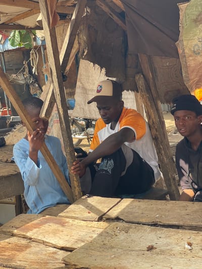 In Chikaji, Nigeria, three individuals are observed amidst a makeshift structure. One person in a light blue shirt supports a wooden beam while looking directly towards the viewer. Another person wearing a white shirt with orange sleeves works above, appearing to secure elements of the structure. A third person wearing a black beanie looks downward. The structure is made of rough wooden planks and fragments of fabric, with a glimpse of a motorcycle visible in the background.