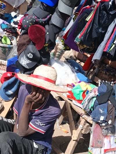 The image depicts an outdoor market stall in Kawaji, Nigeria. A dark-skinned individual is seated in the foreground, wearing a wide-brimmed straw hat with a red band, which obscures their face. They are dressed in a dark blue short-sleeved shirt with white and red trim (a "Paris Saint-Germain" logo is partially visible on the sleeve) and dark trousers. Their right hand is positioned with fingers covering the lower facial area.
The stall prominently displays a variety of merchandise. Numerous baseball caps and other headwear, in colors such as black, grey, blue, maroon, and red, some featuring logos like a Bitcoin "B," "LA," a spider web, and "Adidas," are densely arranged, hanging and stacked. To the right, various garments, including t-shirts, shorts, and bundled socks, are displayed on hangers and a wooden surface. These items include colors such as black, grey, purple, camouflage, and red. Several white plastic bags containing unspecified items are also visible on the wooden display structure.
The background shows additional market elements, including large sacks of granular material, possibly grains, and further cluttered merchandise displays. The ground is dusty and light brown, and the scene is illuminated by strong daylight. The merchandise is presented on makeshift wooden tables and racks.