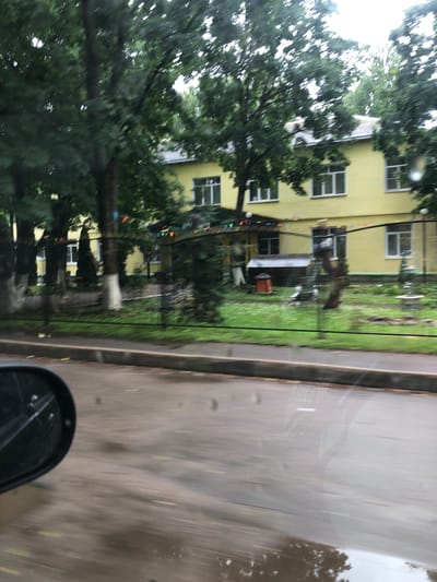 The image shows a blurry, horizontal view from what appears to be inside a vehicle, looking out at a residential street in Safonovo, Russia. The primary subject is a yellow building with multiple windows, possibly an apartment block, flanked by mature trees with lush green foliage. The building has a white trim around the roofline and some of the windows. Below the trees, in front of the building, there's a patch of grass with some scattered items, including what looks like a red object and some colorful flags or bunting strung between poles.
To the right of the building and trees is a wet, grey pavement or road surface, suggesting it has recently rained or is currently raining, as evidenced by water reflections and droplets on what appears to be a car window in the foreground. The overall atmosphere is somewhat muted and overcast, implying it might be daytime but not particularly sunny. The blurry nature of the image suggests movement, likely from the vehicle the photo is being taken from. A small portion of a black car mirror is visible in the bottom right corner, further confirming the perspective. There is no visible text in the image.