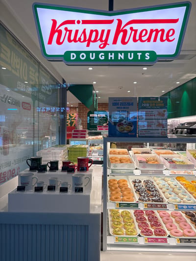 This is a photograph of the exterior of a Krispy Kreme doughnut shop. The shop's iconic green and white sign is prominently displayed above the entrance, reading "Krispy Kreme Doughnuts." Inside, various displays of doughnuts and merchandise are visible through the glass storefront. Several promotional posters are also visible. The image appears to have been taken during the day.