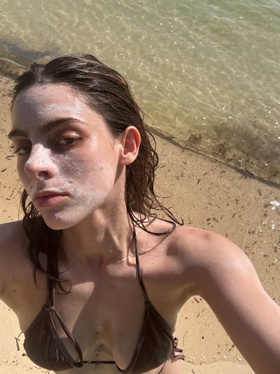A woman in a brown bikini lies on a beach near the water's edge. Her wet, dark hair is tousled around her face, which is covered in white sunscreen. She gazes towards the camera with a serene expression. The clear, shallow water is visible to the left, reflecting sunlight. Tan sand mixed with debris is seen near the water and under her. A tattoo is visible on her torso. The overall image has a sun-kissed, relaxed beach vibe.