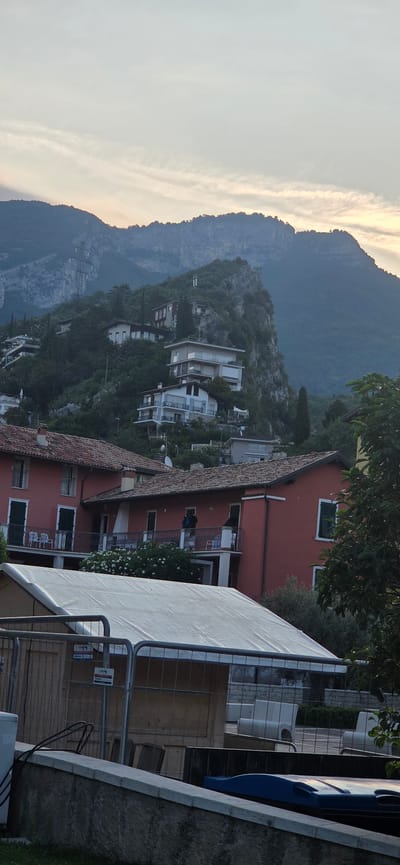 The image captures a view of Torbole, Italy, showcasing a hillside dotted with houses and a backdrop of imposing mountains. In the foreground, a white tent is set up, enclosed by metal fencing. Adjacent to the tent, a stone wall runs horizontally across the image. Beyond the tent and wall, the scene unfolds with a cluster of residential buildings, characterized by their red-tiled roofs and multi-story structures. Some of these buildings are situated on the lower slopes of the hill, while others are perched higher up, nestled amongst dense greenery. The houses vary in architectural style, with some featuring balconies and large windows. The mountainous terrain dominates the background, its rugged slopes partially covered in trees and rock formations. The sky above is a muted gray, with faint clouds hinting at either dawn or dusk. The overall impression is one of a picturesque Italian landscape, combining residential architecture with natural beauty.