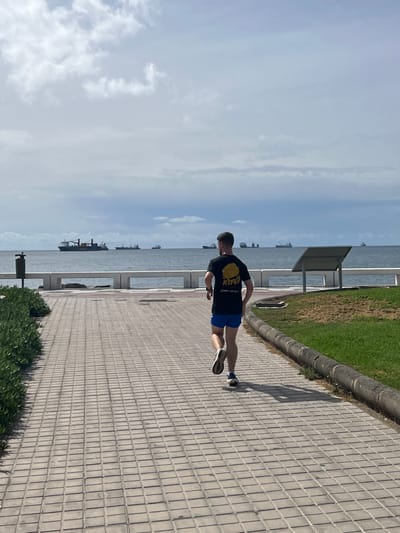 A man in a black t-shirt and blue shorts runs along a tiled sidewalk. He is next to a grassy area bordering the ocean. In the background, several ships are visible on the water. The scene is in Las Palmas, Spain, characterized by the coastal promenade.
