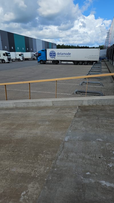 The image depicts an outdoor scene at a logistics facility in Kroņi, Latvia. In the background, a long, multi-colored industrial building is visible, featuring sections in shades of dark gray, light gray, teal, and black. Several trucks are parked in front of the building. The most prominent truck is a blue semi-trailer truck with a white container bearing the "delamode International Logistics" logo. Other white trucks are parked to the left of it. The ground is paved with interlocking gray blocks, marked with white lines indicating parking spaces. A yellow railing with vertical supports runs across the mid-ground. In the foreground, a concrete surface is visible, appearing worn and textured. The sky above is partly cloudy with dramatic cloud formations. The overall environment suggests a commercial logistics or distribution center. There are no people clearly visible in the image.