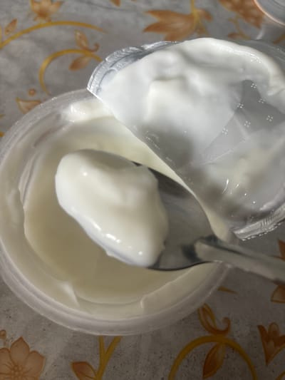 A close-up, overhead shot shows a spoon lifting a dollop of white yogurt from a clear plastic container. The container is open, with a silver foil lid partially peeled back, revealing the creamy contents. The yogurt is smooth and has a glossy sheen. The background features a tablecloth with an ornate floral pattern in shades of beige and orange. The image does not contain any information about the city of Kirov, Russia, or any people.