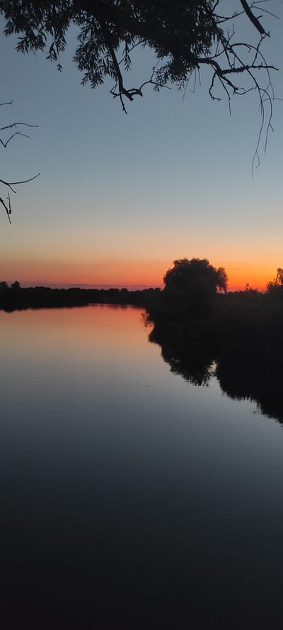This media file depicts a sunset over a body of water, likely a river or lake, in Kobelyachok, Ukraine. The scene is characterized by a dramatic gradient of colors in the sky, transitioning from a soft blue at the top to vibrant oranges and yellows near the horizon, indicative of a sunset.
Dominating the middle of the frame is the dark silhouette of a tree, its branches reaching upwards against the colorful sky. This tree's reflection is clearly visible in the still water below, creating a symmetrical effect. The water itself appears dark and calm, reflecting the colors of the sunset and the surrounding landscape. On the left side of the image, the silhouette of another tree with more intricate branches and foliage is visible against the pale blue sky.
There are no people or man-made objects clearly visible in the image. The overall mood conveyed is one of tranquility and natural beauty, enhanced by the soft light and serene atmosphere of the sunset. The time of day is clearly evening, coinciding with the sunset. No specific weather conditions are apparent beyond the clear sky necessary for a sunset. The location context of Kobelyachok, Ukraine, provides geographical information for the scene. There is no visible text in the image.